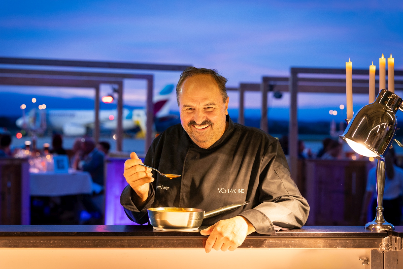 Koche mit Pfanne auf Theke, dahinter Flughafen im Dämmerlicht