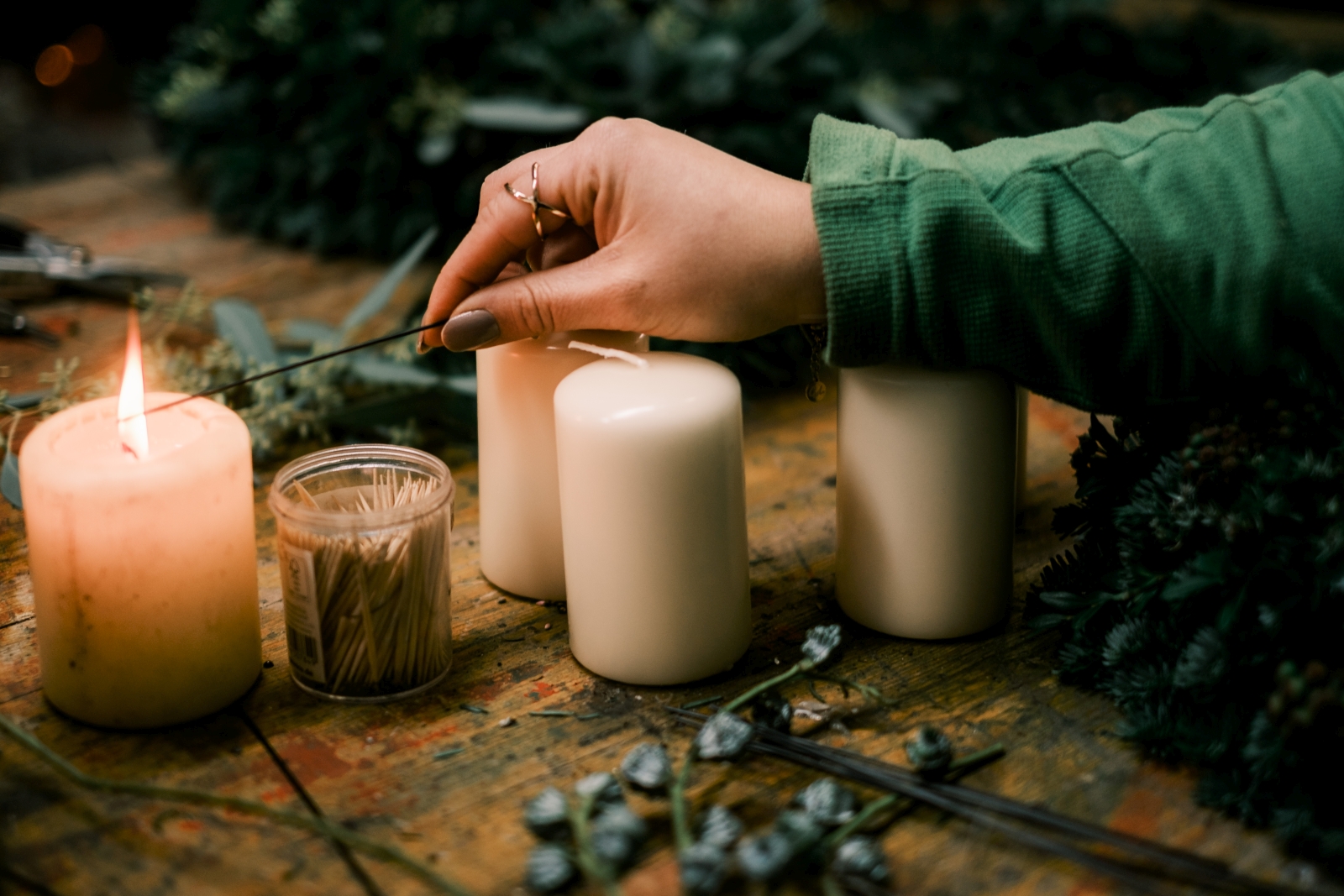 mehrere Adventkranzkerzen auf Holztisch, rundherum Material für Adventkranz; eine Kerze wird gerade angezunden