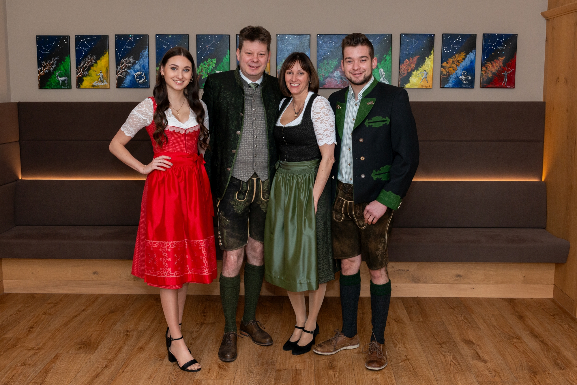 Vier Personen in Tracht schauen in die Kamera, zwei Frauen, zwei Männer, dahinter eine braune Bank und viele Bilder an der Wand