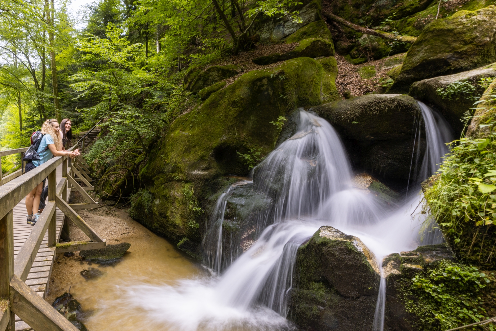 Zwei Frauen mit Rucksack auf einer Holzbrücke mit Blick auf einen Wasserfall