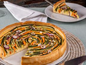 Angeschnitte Tarte mit Zucchini und Kürbis in dünnen Scheiben in Spiralen belegt. Darunter weißer Teller und beiges Tischset, im Hintergrund ein Tortenstück der Tarte.