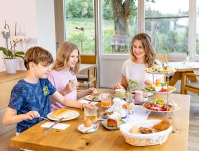 Frau und zwei Kinder am Frühstückstischmit Speisen auf Etagere, Brotkorb, Eiern, Säften und Kuchen, dahinter große Fenster mit Blick in einen Garten