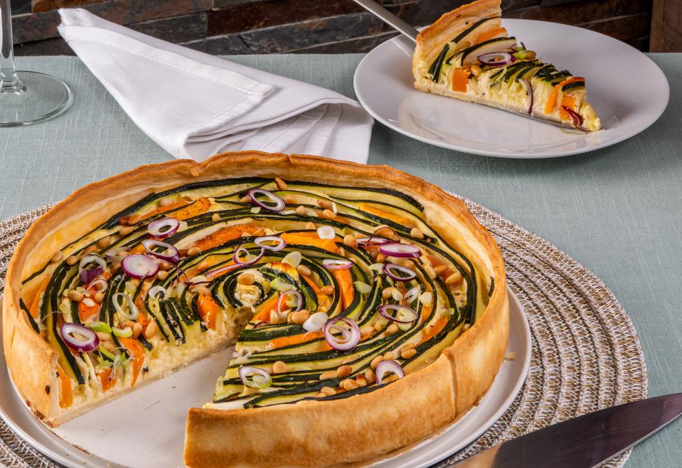 Angeschnitte Tarte mit Zucchini und Kürbis in dünnen Scheiben in Spiralen belegt. Darunter weißer Teller und beiges Tischset, im Hintergrund ein Tortenstück der Tarte.