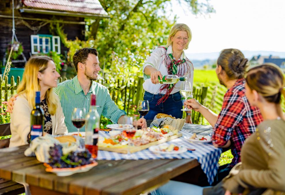 Fröhliche Menschen sitzen um einen Holztisch mit Jause und Wein, Frau schenkt Wein ein