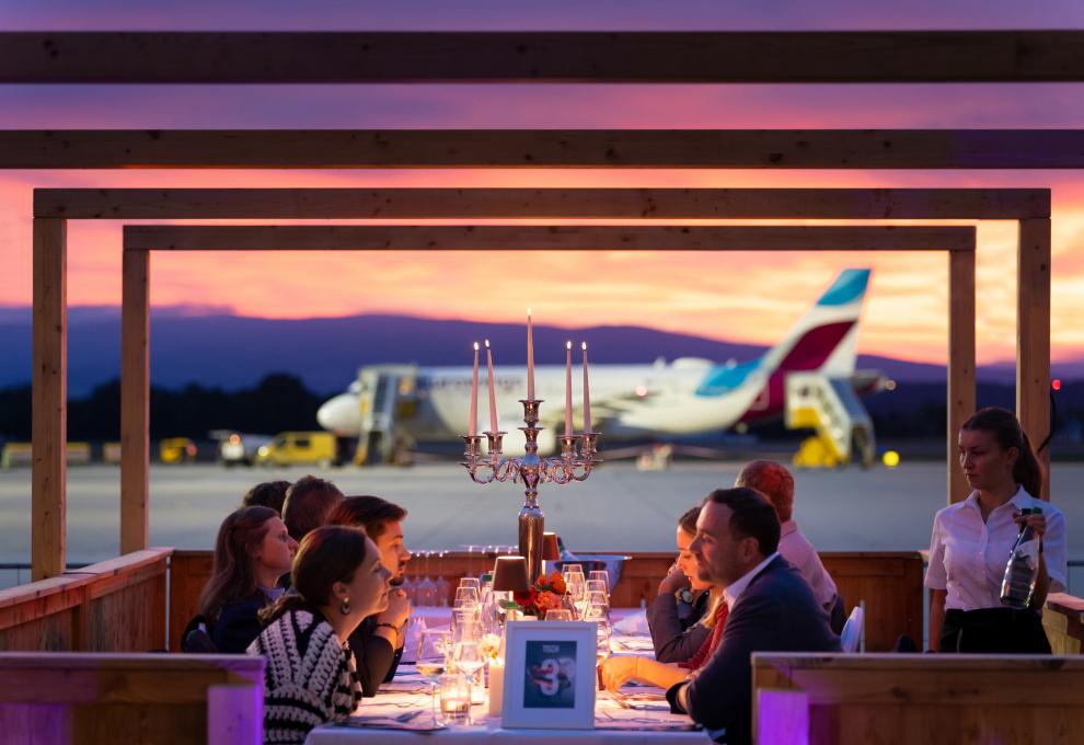 Menschen sitzen am festlich gedeckten Tisch, dahinter ein Flugzeug am Flughafen