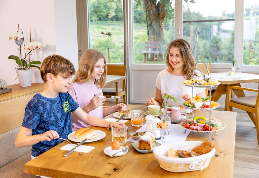 Frau und zwei Kinder am Frühstückstischmit Speisen auf Etagere, Brotkorb, Eiern, Säften und Kuchen, dahinter große Fenster mit Blick in einen Garten