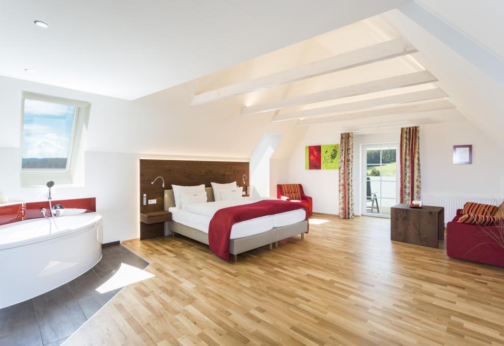 Hotelzimmer mit Doppelbett, großen Fenstern und Badewanne im Zimmer. Farben: weiß, rot, Holzboden