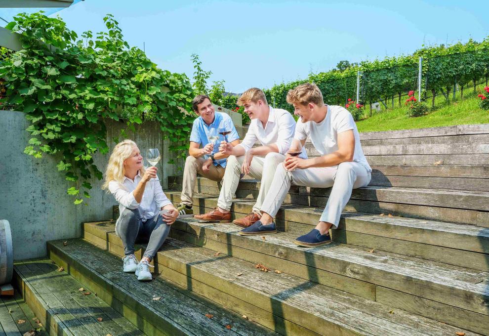 Eine Frau und drei Männer sitzen auf einer Holztreppe, im Hintergrund Weinberge