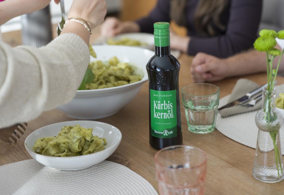 Szene am Tisch mit Bandnudeln in großer Schüssel, Wassergläsern und einer Flasche Steiriches Kürbiskernöl g.g.A. von Steirerkraft